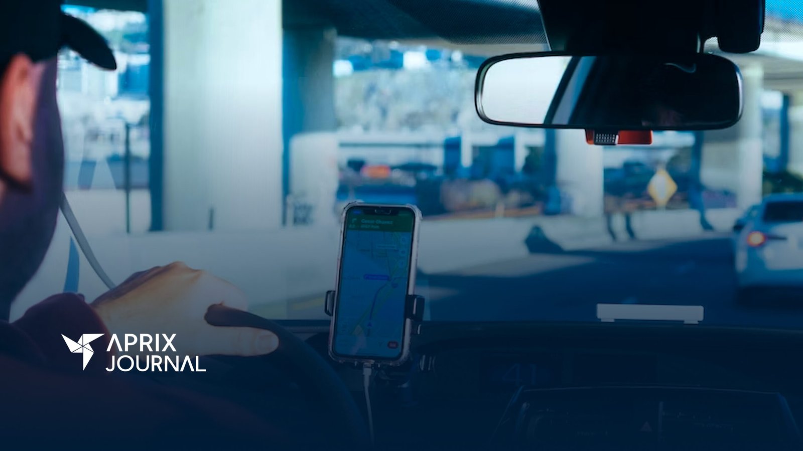 A foto mostra a visão do banco de trás de um carro. É possível ver a lateral do rosto do motorista, com uma mão ao volante, e, ao lado do volante, um celular com a tela mostrando um aplicativo de trânsito