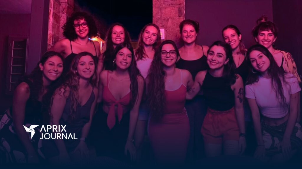 Um grupo de mulheres enfileiradas e abraçadas sorriem em direção À CÂMERA, EM UM AMBIENTE ILUMINADO COM LUZES ROSA