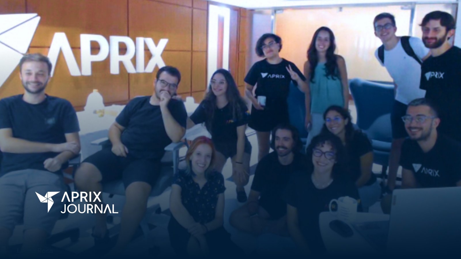 Um grupo de pessoas está em uma sala com uma placa da Aprix ao fundo. Algumas pessoas estão em pé, outras sentadas em sofás e cadeiras, ou no chão. O grupo está descontraído e sorri em direção à câmera
