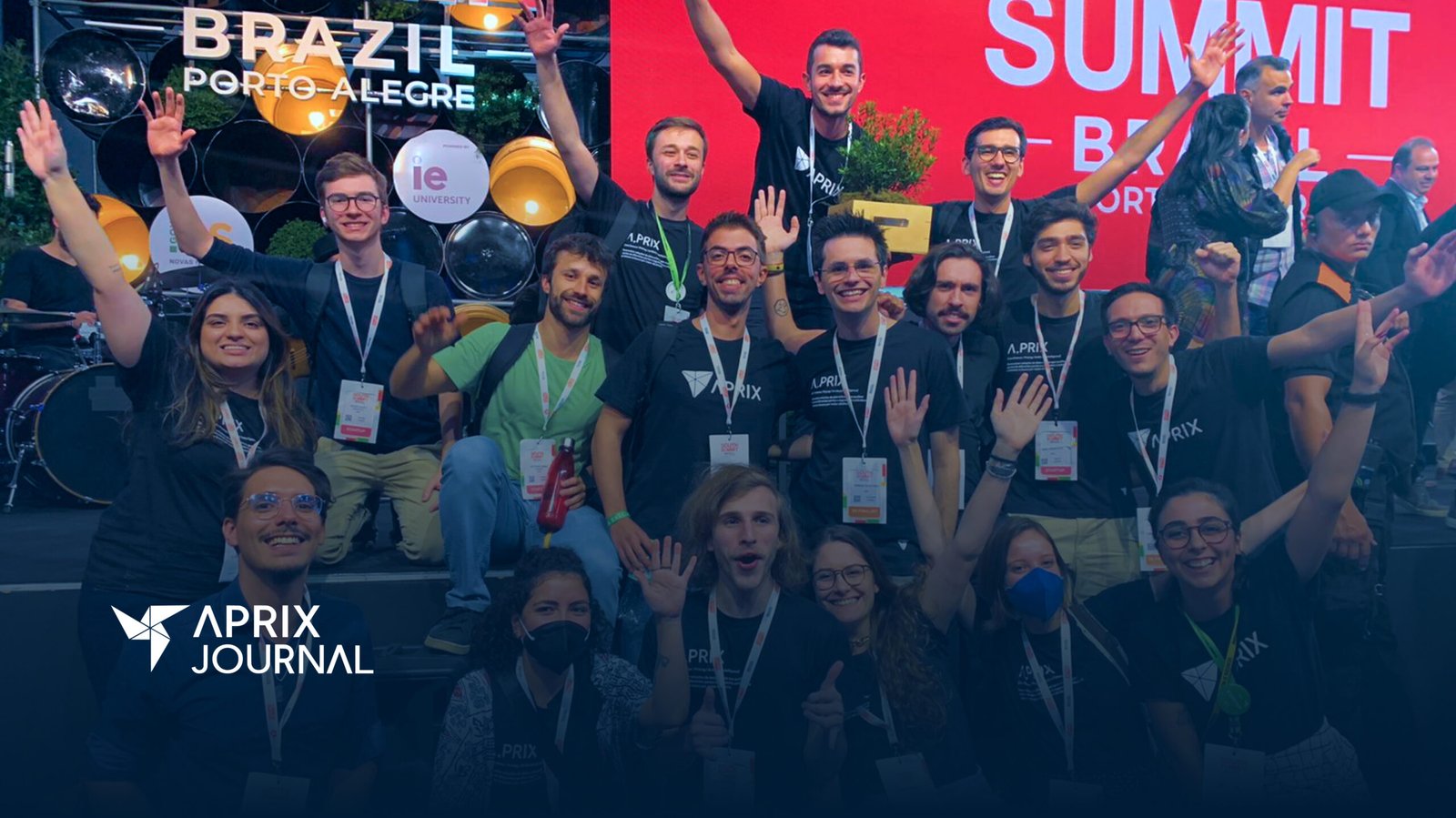 Um grupo de pessoas, muitas vestindo camisetas com o logo da Aprix, sorriem e comemoram à frente de um palco. Em uma tela, ao fundo, há o logo do South Summit Brazil 2023