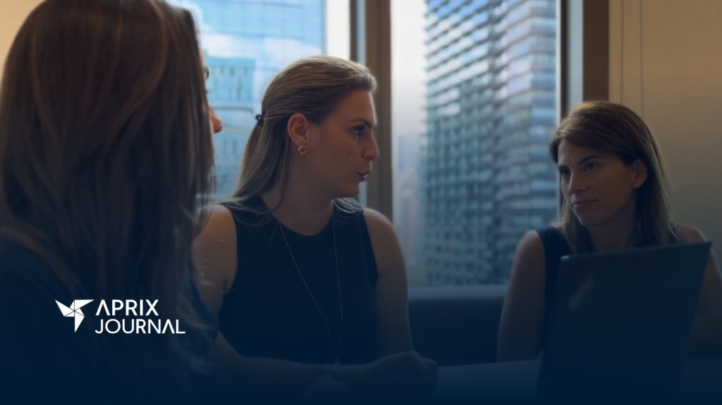 Três mulheres em uma reunião em um ambiente corporativo, com um grande janelão ao fundo revelando prédios modernos. Duas delas estão de frente para a câmera, enquanto a terceira aparece de costas. A mulher ao centro fala enquanto a outra escuta atentamente, com um laptop aberto à sua frente. O logotipo 'Aprix Journal' está posicionado no canto inferior esquerdo da imagem.